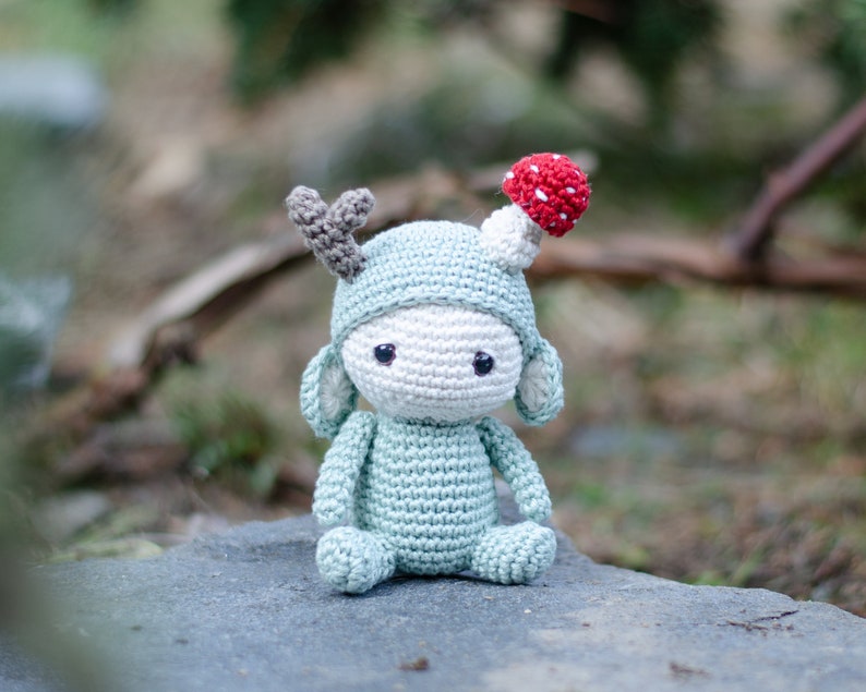 May include: A handmade crochet doll in a light blue outfit with a matching hat and gray antlers. The doll has a white face and a red and white mushroom on top of the hat. The doll is sitting on a gray stone.