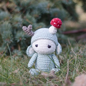 May include: A handmade crochet doll in a whimsical forest theme. The doll is primarily light blue with a white face and a mushroom on its head. It has gray antlers and is sitting in the grass.