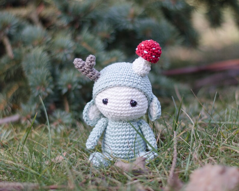 May include: A handmade crochet doll in a whimsical forest theme. The doll is primarily light blue with a white face and a mushroom on its head. It has gray antlers and is sitting in the grass.