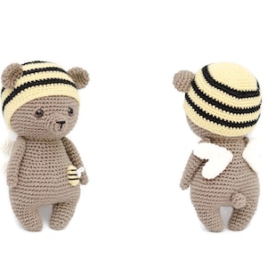 Pode incluir: Um urso de pelúcia de crochê usando um chapéu listrado com tema de abelha e pequenas asas brancas. O urso tem uma pequena aplicação de abelha na barriga. O urso é marrom com um chapéu listrado amarelo claro e preto.