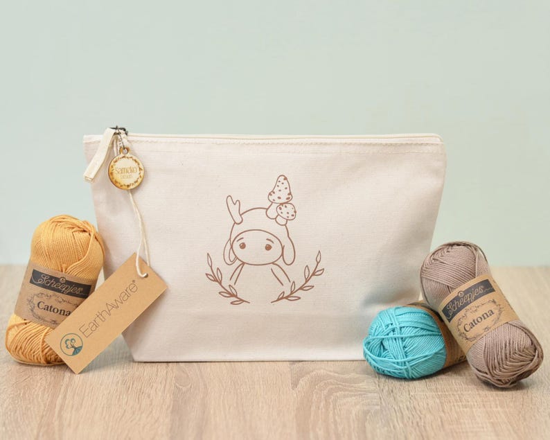 Könnte beinhalten: Eine cremefarbene Canvas-Tasche mit einer Pilz-Charakter-Illustration, einem Reißverschluss und einem Holzanhänger. Die Tasche ist von Garnknäueln in Gelb, Türkis und Taupe umgeben, mit Scheepjes Catona-Etiketten und einem EarthAware-Anhänger.