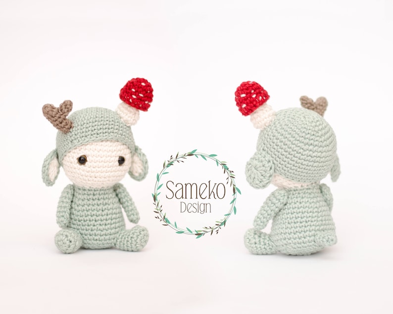 May include: Crocheted deer doll with a mushroom hat. The doll is light green with a cream-colored face and ears. It has brown antlers and a red and white mushroom on its head. The doll is sitting with its arms and legs outstretched.
