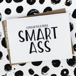 May include: A white card with black text that reads "Congratulations Smart Ass" on a white background with black paint splatters.
