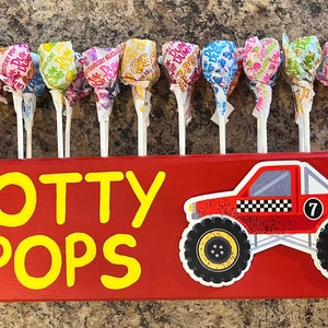 May include: A red wooden sign with the words "POTTY POPS" in yellow letters. A red monster truck with a black checkered flag and the number "7" is on the right side of the sign. There are 18 colorful lollipops in a row above the sign.