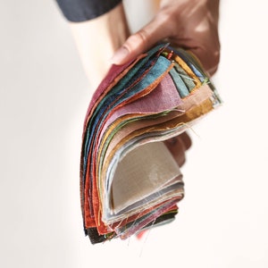 Puede incluir: Una mano sostiene un abanico de muestras de tela en varios colores. Las muestras incluyen tonos de rojo, verde, azul, amarillo y tonos neutros. La tela parece ser lino o un material similar, con una superficie ligeramente texturizada.