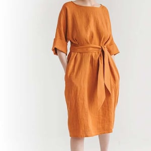 May include: An orange linen dress with a boat neck, elbow-length sleeves, and a matching waist tie. The dress has a knee-length skirt and pockets. The model is standing in front of a white background.