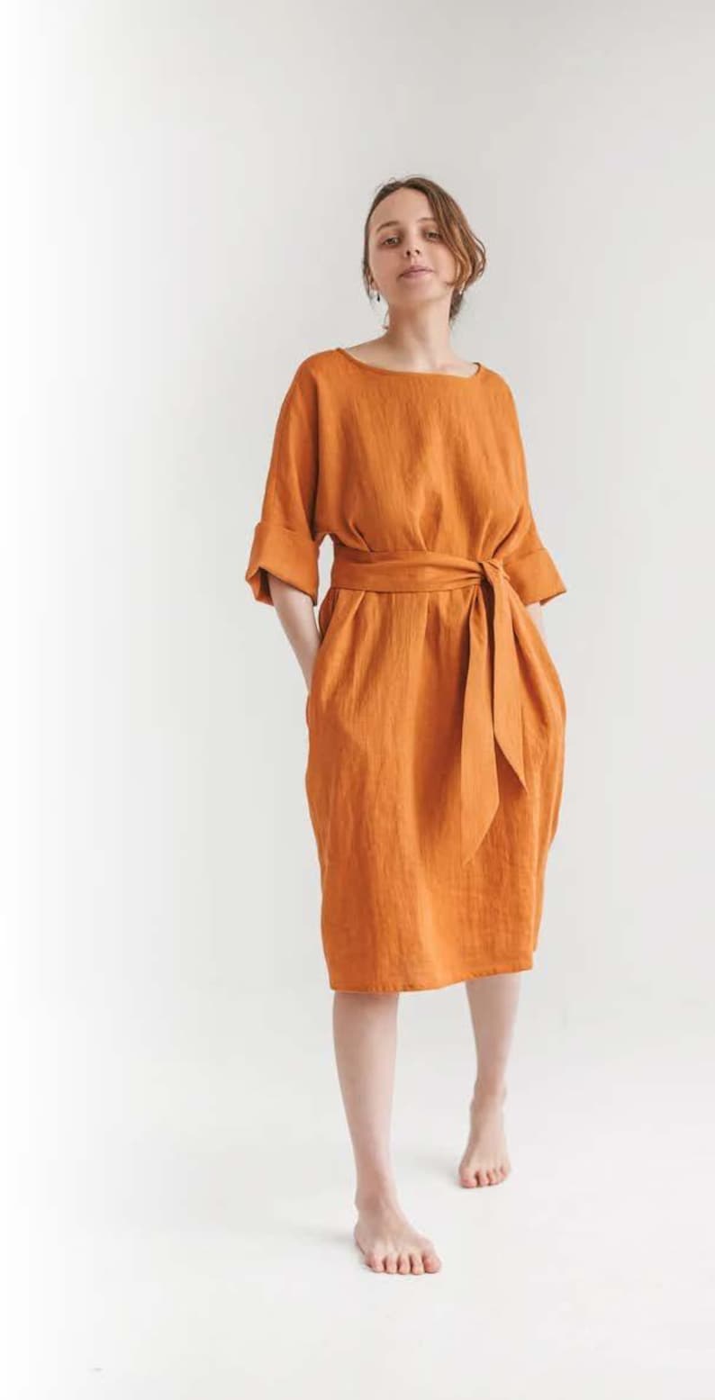 May include: An orange linen dress with a boat neck, elbow-length sleeves, and a matching waist tie. The dress has a knee-length skirt and pockets. The model is standing in front of a white background.