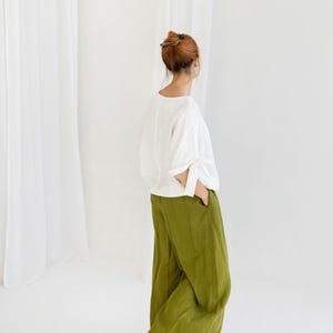 K&ouml;nnte beinhalten: Eine Person tr&auml;gt ein wei&szlig;es Langarm-Oberteil und eine olivgr&uuml;ne Hose mit weitem Bein. Das Outfit ist schlicht und elegant, mit Fokus auf Komfort und Stil. Der Hintergrund ist hellwei&szlig;.