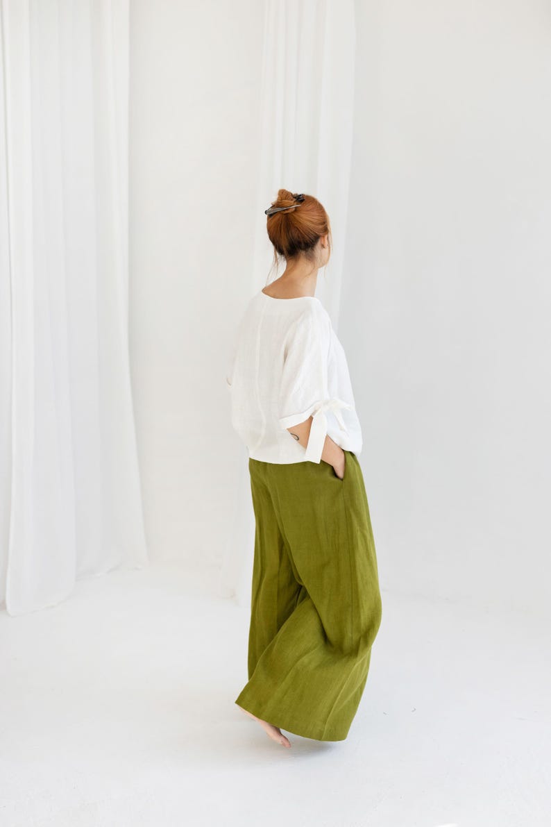 K&ouml;nnte beinhalten: Eine Person tr&auml;gt ein wei&szlig;es Langarm-Oberteil und eine olivgr&uuml;ne Hose mit weitem Bein. Das Outfit ist schlicht und elegant, mit Fokus auf Komfort und Stil. Der Hintergrund ist hellwei&szlig;.