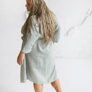May include: A light green linen robe with a tie waist. The robe has three-quarter sleeves and falls to the mid-calf. The model is standing in front of a white textured wall.