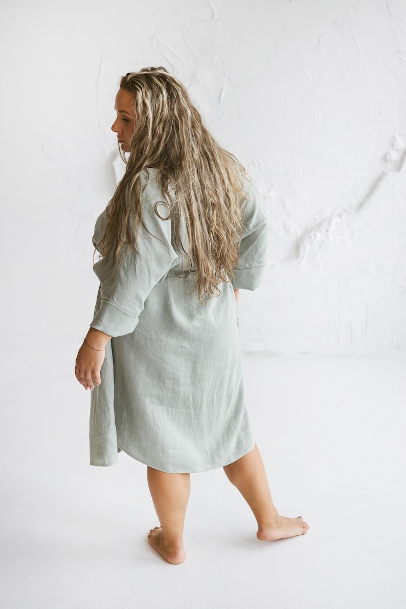 May include: A light green linen robe with a tie waist. The robe has three-quarter sleeves and falls to the mid-calf. The model is standing in front of a white textured wall.
