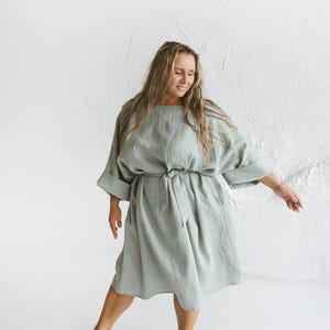 May include: A light green linen dress with a round neckline, elbow-length sleeves, and a tie waist. The dress falls to the mid-calf and has pockets. The model is wearing the dress in a studio setting with a white background.
