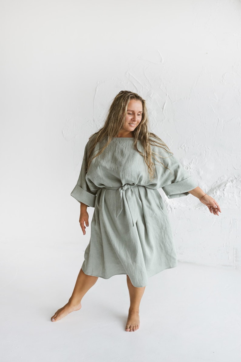 May include: A light green linen dress with a round neckline, elbow-length sleeves, and a tie waist. The dress falls to the mid-calf and has pockets. The model is wearing the dress in a studio setting with a white background.