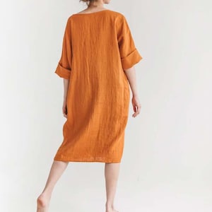 May include: An orange linen dress with a relaxed fit and elbow-length sleeves. The dress falls to the mid-calf and has a simple, minimalist design. The model is standing barefoot against a white background.