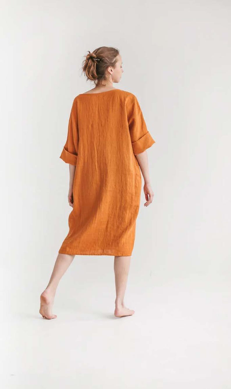May include: An orange linen dress with a relaxed fit and elbow-length sleeves. The dress falls to the mid-calf and has a simple, minimalist design. The model is standing barefoot against a white background.