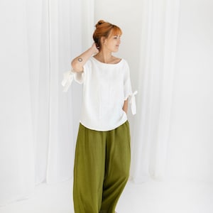 May include: A person wearing a white linen top with bow details on the sleeves and olive green wide-leg trousers. The outfit is simple and elegant, suitable for casual wear. The background is plain white.