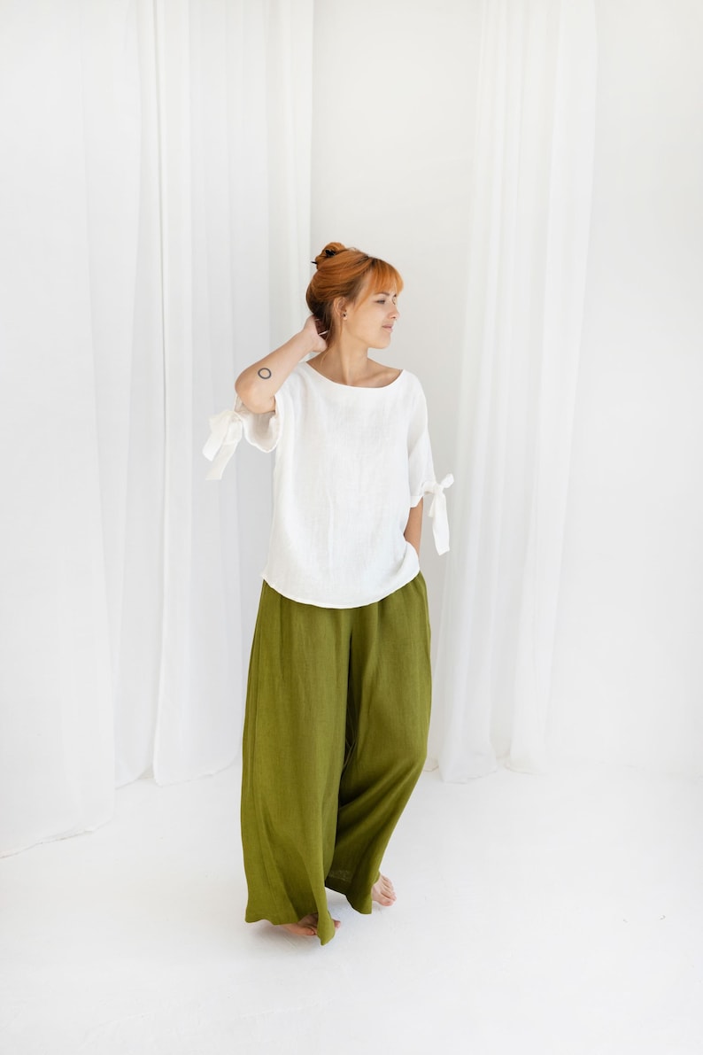 May include: A person wearing a white linen top with bow details on the sleeves and olive green wide-leg trousers. The outfit is simple and elegant, suitable for casual wear. The background is plain white.