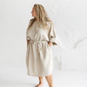 May include: A beige linen dress with a round neckline, elbow-length sleeves, and a tie waist. The dress has pockets and falls to the mid-calf. The model is standing in front of a white textured wall.