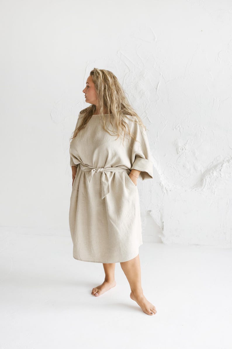 May include: A beige linen dress with a round neckline, elbow-length sleeves, and a tie waist. The dress has pockets and falls to the mid-calf. The model is standing in front of a white textured wall.
