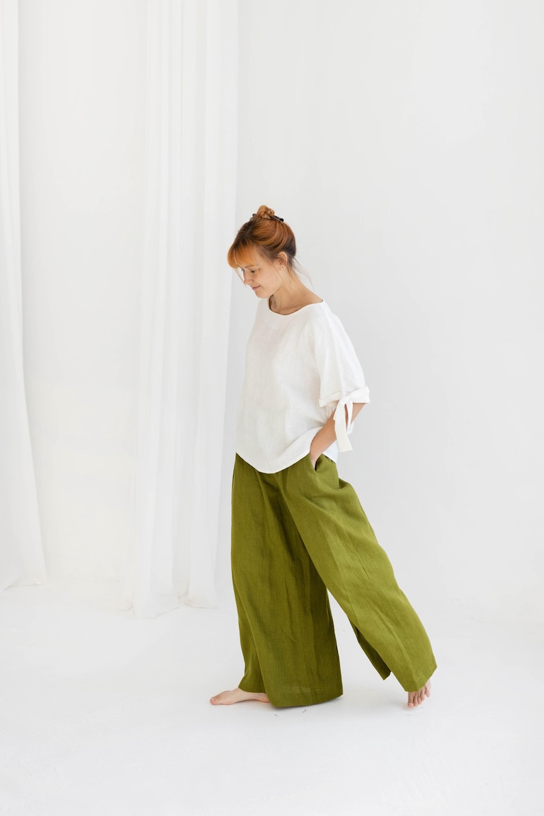 K&ouml;nnte beinhalten: Eine Person tr&auml;gt ein wei&szlig;es Leinenoberteil mit Bindeb&auml;ndern an den &Auml;rmeln und eine olivgr&uuml;ne Leinenhose. Das Outfit ist schlicht und elegant, mit einer lockeren Passform. Der Hintergrund ist eine schlichte wei&szlig;e Wand und Vorh&auml;nge.