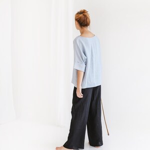 May include: A person wearing a light blue top with rolled sleeves and black wide-leg pants. The outfit is simple and minimalist, with a focus on comfort and style. The clothing is made of a soft, flowing fabric.