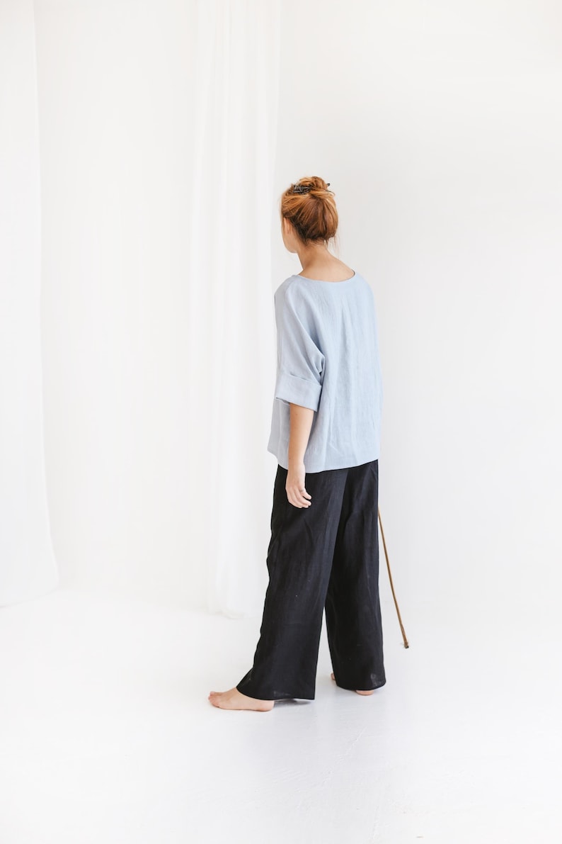 May include: A person wearing a light blue top with rolled sleeves and black wide-leg pants. The outfit is simple and minimalist, with a focus on comfort and style. The clothing is made of a soft, flowing fabric.
