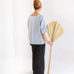 May include: A person wearing a light blue linen top and black linen pants. The person is holding a large, dried palm leaf fan with a wooden handle. The background is a plain white wall and floor.