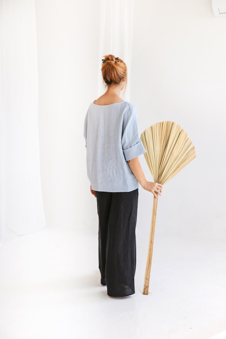 May include: A person wearing a light blue linen top and black linen pants. The person is holding a large, dried palm leaf fan with a wooden handle. The background is a plain white wall and floor.