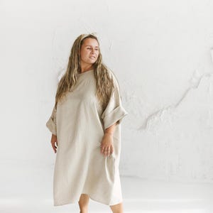 May include: A beige linen dress with a loose, flowing silhouette. The dress features a round neckline, wide sleeves, and a knee-length hem. The model is wearing a headband and is standing in front of a white wall.