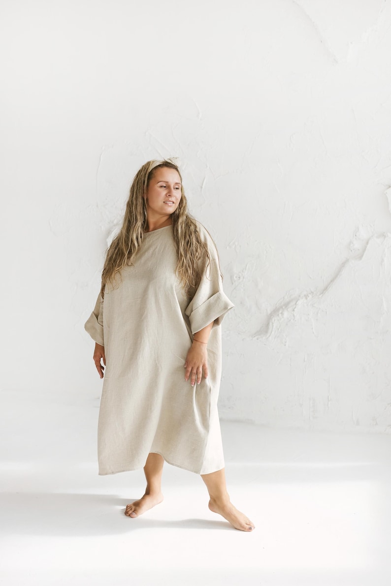 May include: A beige linen dress with a loose, flowing silhouette. The dress features a round neckline, wide sleeves, and a knee-length hem. The model is wearing a headband and is standing in front of a white wall.