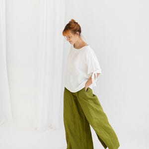 May include: A woman in a white top with tie details on the sleeves and olive green wide-leg trousers. The outfit is made of a lightweight fabric. The background is plain white. The trousers are a vibrant shade of green.