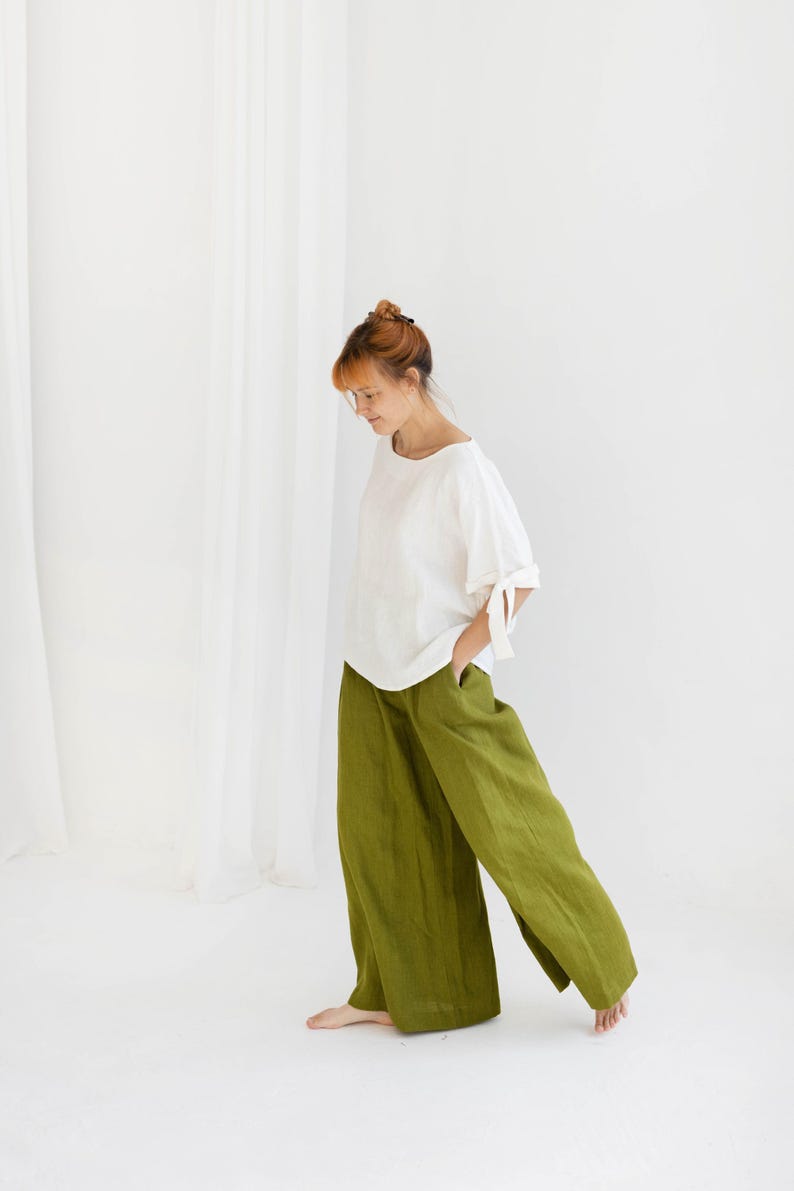 May include: A woman in a white top with tie details on the sleeves and olive green wide-leg trousers. The outfit is made of a lightweight fabric. The background is plain white. The trousers are a vibrant shade of green.
