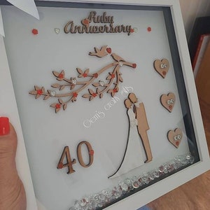 Könnte beinhalten: Ein weißer Rahmen mit weißem Hintergrund und einem Holzbaum mit roten Blüten und Blättern. Der Baum hat zwei Vögel auf einem Ast. Die Worte "Ruby Anniversary" sind in braunen Buchstaben geschrieben. Die Zahl "40" ist in braunen Buchstaben geschrieben. In der oberen rechten Ecke befinden sich drei rote Herzen mit weißen Punkten. Die Worte "Gem's crafty gifts" sind in weißen Buchstaben geschrieben.