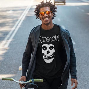 K&ouml;nnte beinhalten: Ein Mann tr&auml;gt ein schwarzes T-Shirt mit einem wei&szlig;en Totenkopf-Motiv und dem aufgedruckten Text "MAROON 5". Er tr&auml;gt eine schwarze Jacke und eine Sonnenbrille.