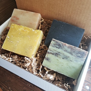 May include: A gift box filled with four bars of handmade soap. The soaps are various colors, including yellow, black, and green, and are nestled in brown paper filler. The box is white and sits on a wooden surface.