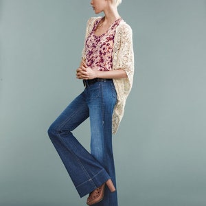 May include: A woman wearing a white crocheted cardigan over a floral print tank top and blue denim flared jeans. She is wearing brown platform shoes.