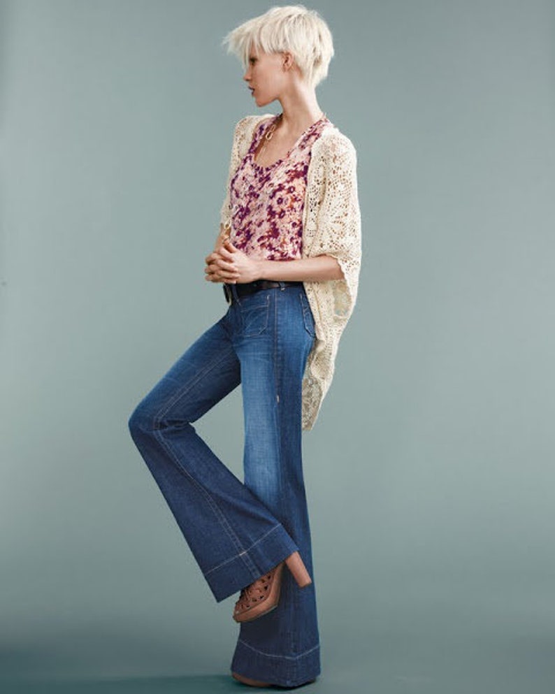 May include: A woman wearing a white crocheted cardigan over a floral print tank top and blue denim flared jeans. She is wearing brown platform shoes.