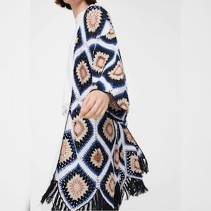 May include: A white crochet cardigan with a geometric pattern in shades of blue, black, and brown. The cardigan has a fringe trim.