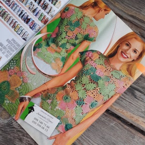 May include: A close-up of a crocheted top with a floral design in shades of green, pink, and beige. The top has short sleeves and a round neckline. The model is wearing the top and smiling.