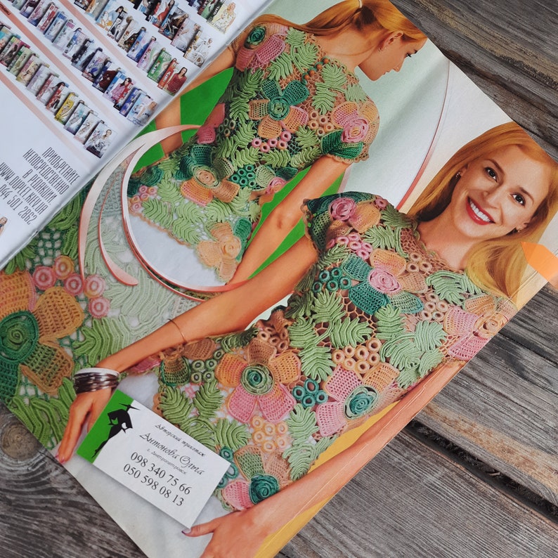 May include: A close-up of a crocheted top with a floral design in shades of green, pink, and beige. The top has short sleeves and a round neckline. The model is wearing the top and smiling.