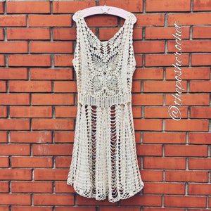 May include: A white crochet dress with a delicate pattern hangs on a pink hanger against a brick wall. The dress has a sleeveless design and a flowing skirt.