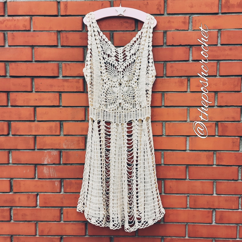 May include: A white crochet dress with a delicate pattern hangs on a pink hanger against a brick wall. The dress has a sleeveless design and a flowing skirt.