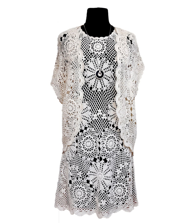 May include: A white crocheted dress and matching cardigan with a delicate floral pattern. The dress has a fitted bodice and a flowing skirt. The cardigan has a short, cropped style.