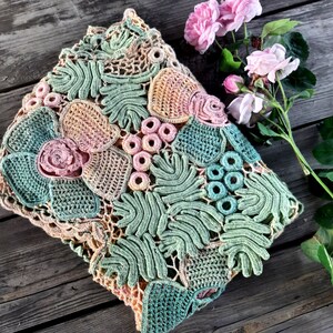 May include: A crocheted pillow with a floral design in shades of green, pink, and beige. The pillow is made with intricate stitches and has a delicate, feminine feel.