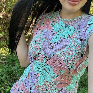 May include: A crocheted top with a floral design in shades of pink, purple, and green. The top is sleeveless and has a round neckline.