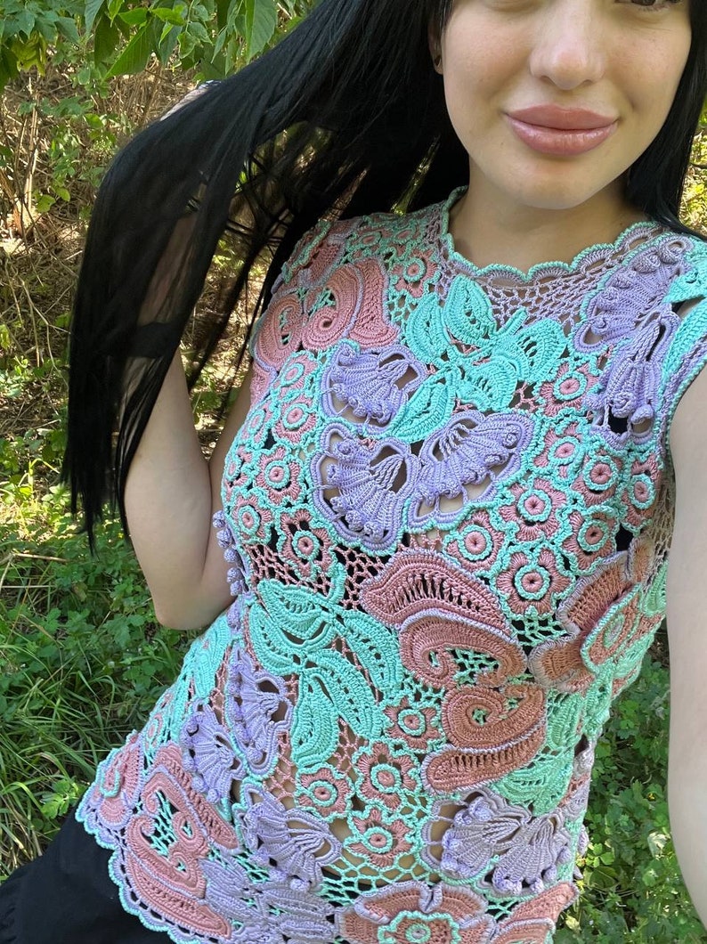 May include: A crocheted top with a floral design in shades of pink, purple, and green. The top is sleeveless and has a round neckline.