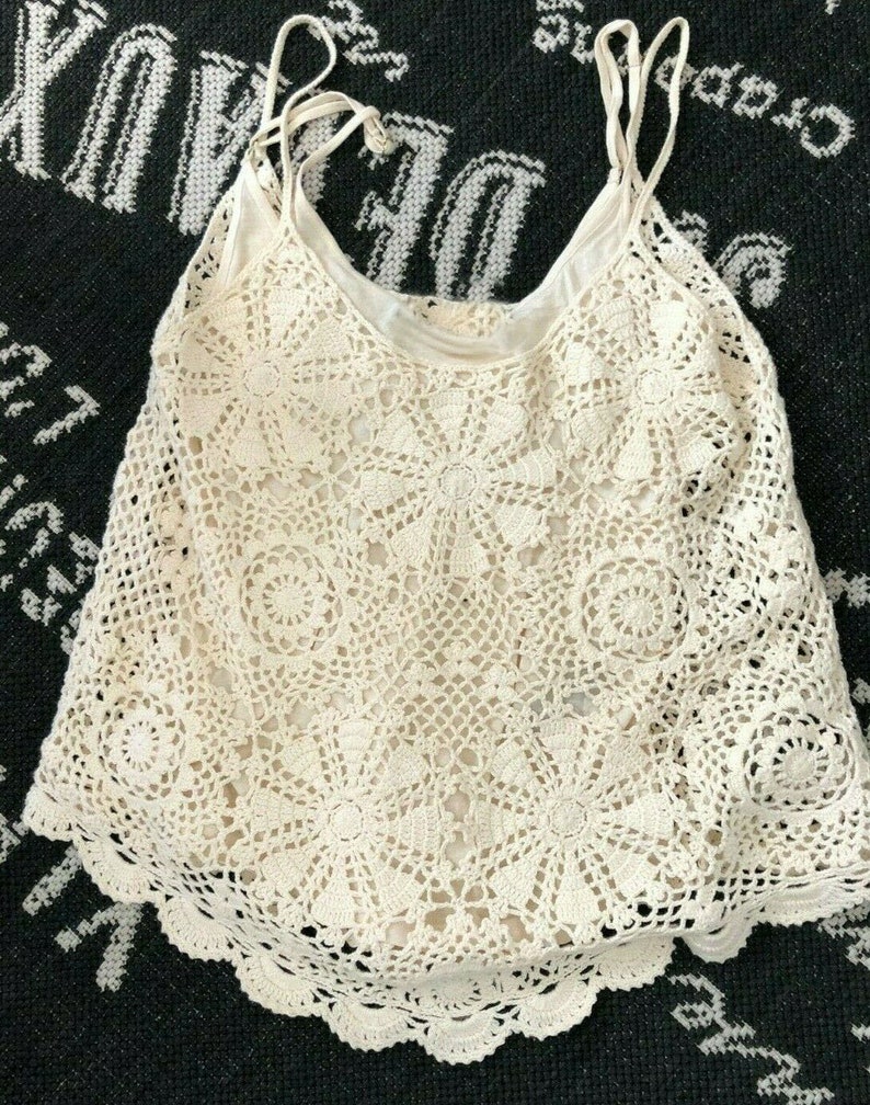 May include: A white crochet tank top with a delicate floral pattern. The top has thin straps and a scalloped hem.