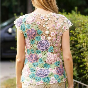 May include: A white crochet top with a floral design featuring pink, purple, and green roses. The top has a delicate, openwork pattern and is sleeveless.