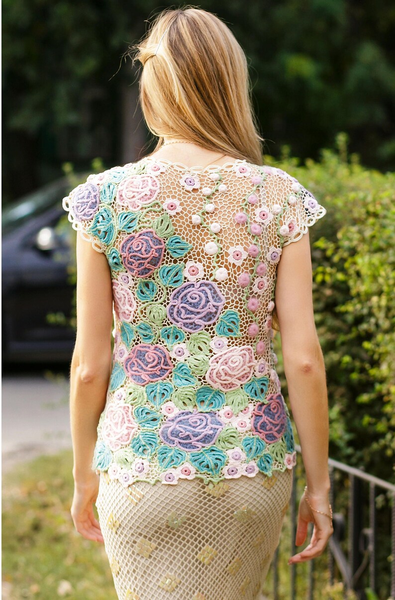 May include: A white crochet top with a floral design featuring pink, purple, and green roses. The top has a delicate, openwork pattern and is sleeveless.