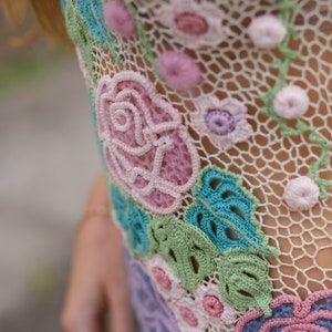 May include: Close-up of a crocheted garment with a floral design. The garment is made of a delicate, white lace with pink, blue, and green flowers. The flowers are intricately crocheted and have a three-dimensional appearance.